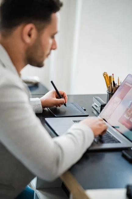 A photographer working at a creative setup with a laptop and Wacom tablet, focused on editing photos indoors.